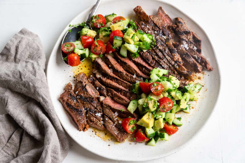 A round white plate showing sliced steak and cucumber and avocado salad, with a grey linen serviette.