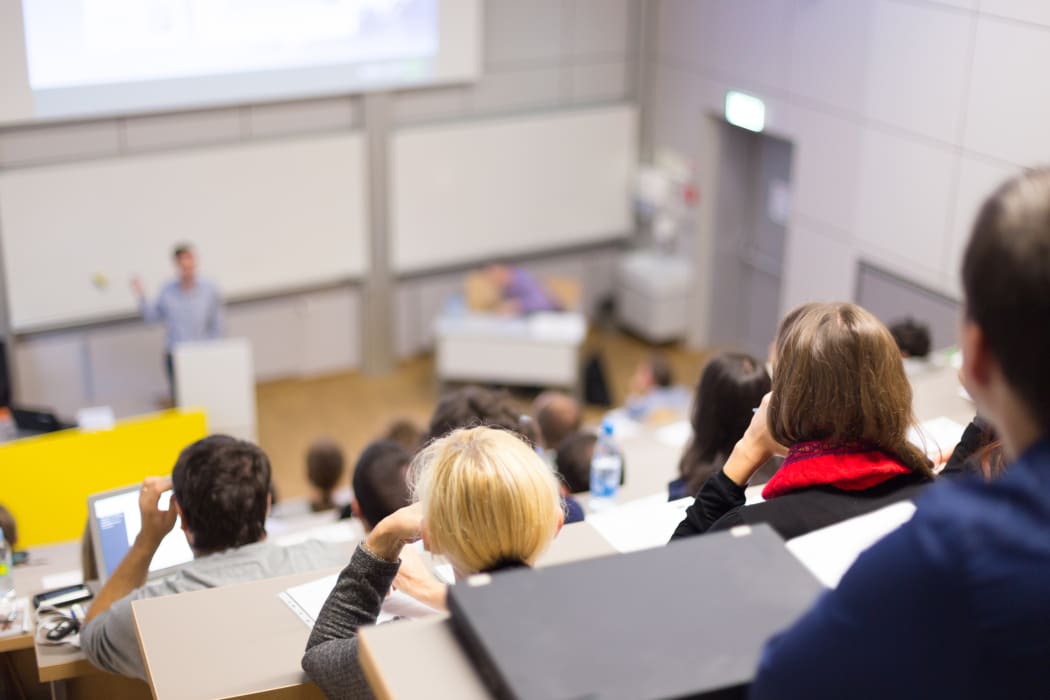 Professor giving presentation in lecture hall at university. Participants listening to lecture and making notes.