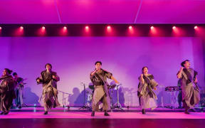 Five performers on a stage lit up in pink lights. Behind them is a live band kit. They are mid-motion, dancing and singing.