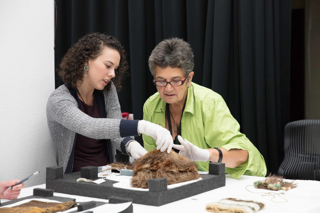 Christina Wirihana and Kate Blair closely inspecting kete muka (flax fibre bag) made with kiwi and kākā feathers.
