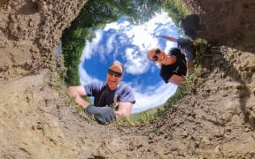 Kirstin and Matt peer into a hole in the ground, smiling.