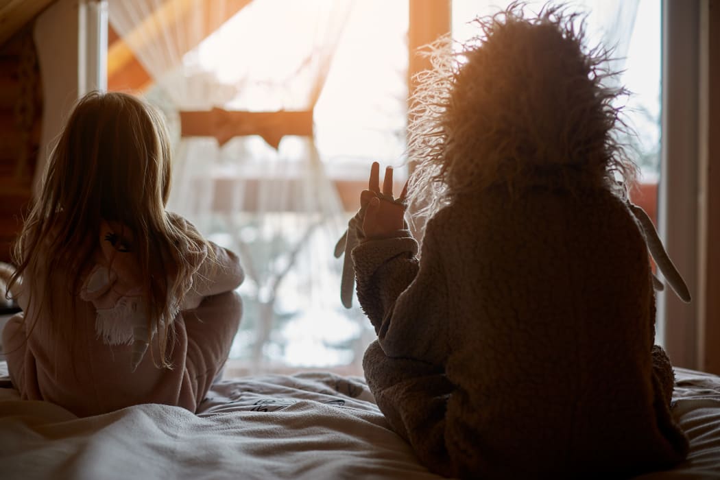 Back view of kids in warm costumes sitting together on bed in warm sunlight
