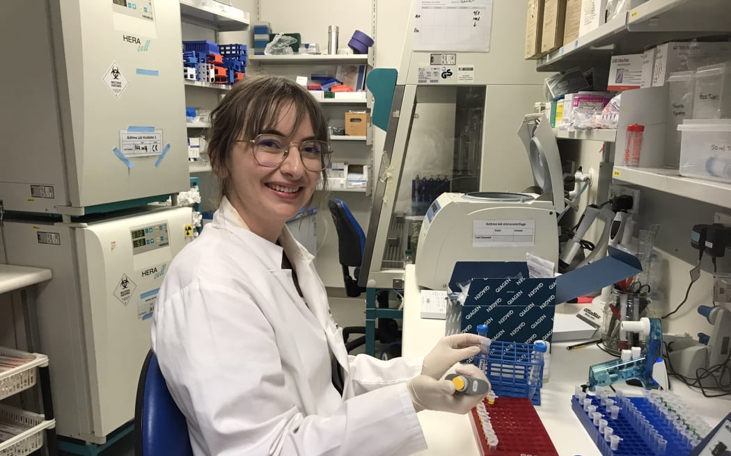 A woman in a white lab coat sits at a lab bench holding a pipette with a row of eppendorf tubes in a red holder and a tray of pipette tips open. The background is crowded with scientific equipment and a fume hood.