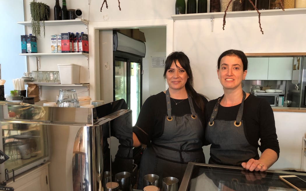Two women, Keita Powley and Siza Avakh, stand side by side behind the counter in a coffee shop. They are wearing black tops and aprons.