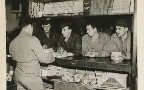 An old photograph. Four US Marines sit at a bar bench, being served by a clerk. They all wear fatigues and hats.
