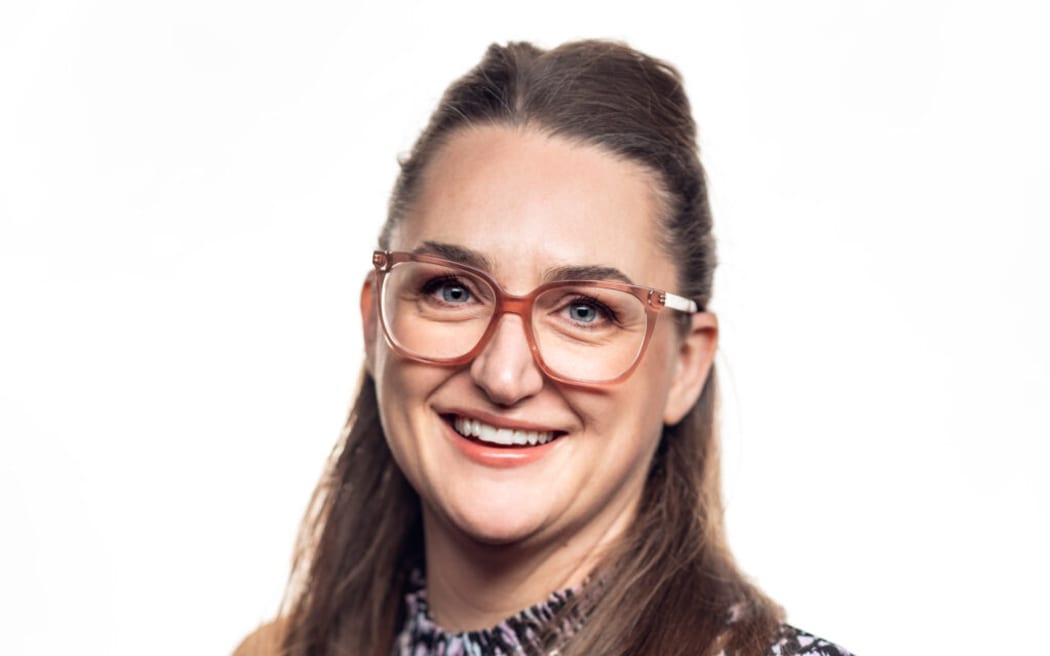 Alice smiles at the camera. She wears a black and white patterned shirt and glasses.