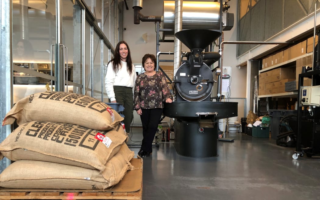 Two women, Steph Noble and Roz Cattell, stand in a coffee roastery room. There are sacks of coffee beans in the foreground, and a big metal machine in the centre of the room. Boxes line the walls. The woman on the left is tall, and wearing a white long sleeved top and black pants. The woman on the right is shorter, wearing a black floral top and black pants.