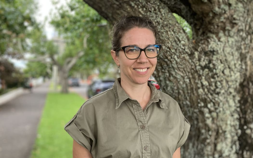 Erika smiles at the camera. She is wearing a khaki short-sleeved shirt. She is standing in front of a large tree on the berm of a residential street.