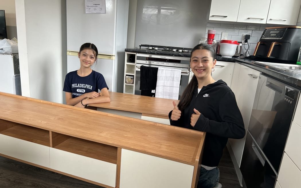 Picture of two young women smiling at camera, sitting among furniture pieces.
