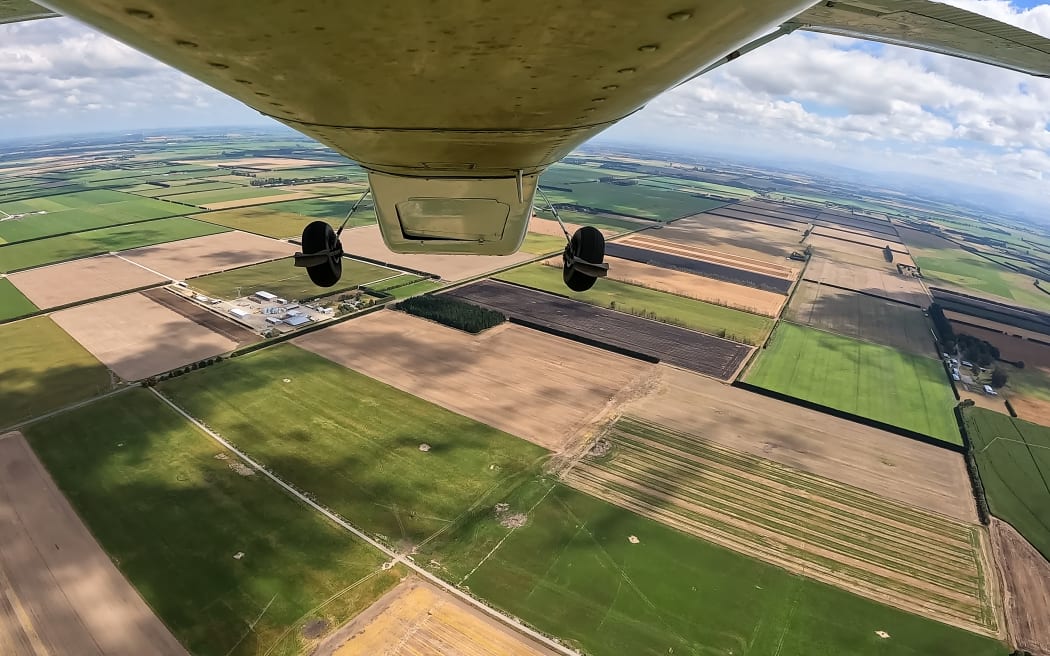Aerial view from underneath an airplane showing a patchwork of agricultural fields in different shades of green and brown. The horizon is visible in the distance under a partly cloudy sky.