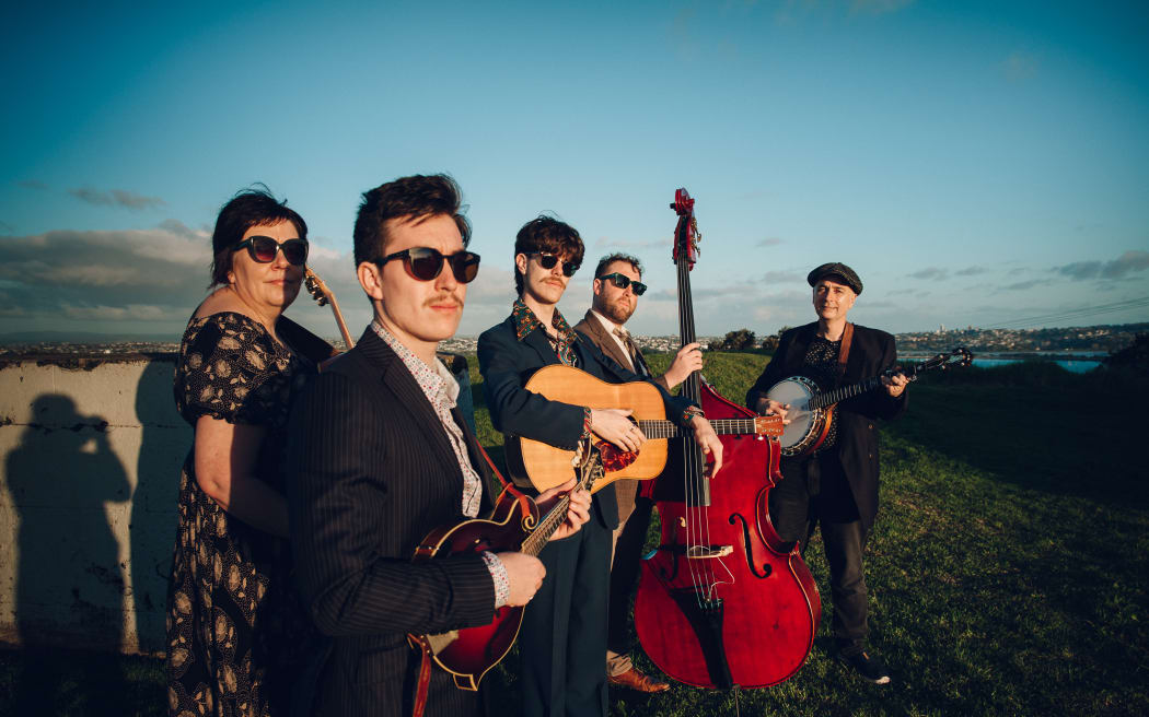 This is a phoyo of the band in a field. They are stood in a group and holding their instruments. All of them are wearing sunglasses exceot the banjo player. That probably makes sense practically as he is facing a different direction to the other four.