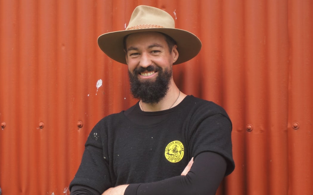 A man wearing a black long-sleeved shirt and a tan-coloured wide-brimmed hat stands with his arms folded in front of burnt orange corrugated iron. He has a dark beard and is smiling warmly.