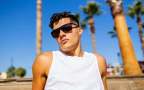 A man in sunglasses and a white tank top pouts at the camera with a blue sky in the background.