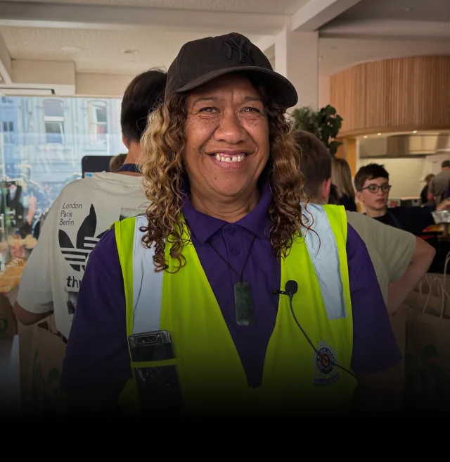 Māori warden Marino Paikea smiles at the camera. A group of people surround her, packing food into brown shopping bags. Marino Paikea is wearing a yellow hi-vis vest.