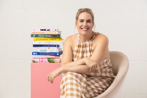 An image of Christchurch-based food writer and chef Sam Parish. Sam is wearing a brown and white checked jumpsuit and sitting next to a stack of cookbooks.
