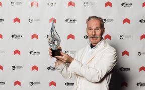 DineAid founder and award-winning chef Mark Gregory is inducted into the Resturant Association Hall Of Fame. He is wearing a white jacket, in the photo it looks a bit like a lab coat. This is probably because Mark looks a bit like a stereotypical "mad professor" with a grey moustache. He is stood in front of a backdrop featuring the awards logo and holding his trophy. The trophy is a kind of teardrop shaped metal object with two holes in it.