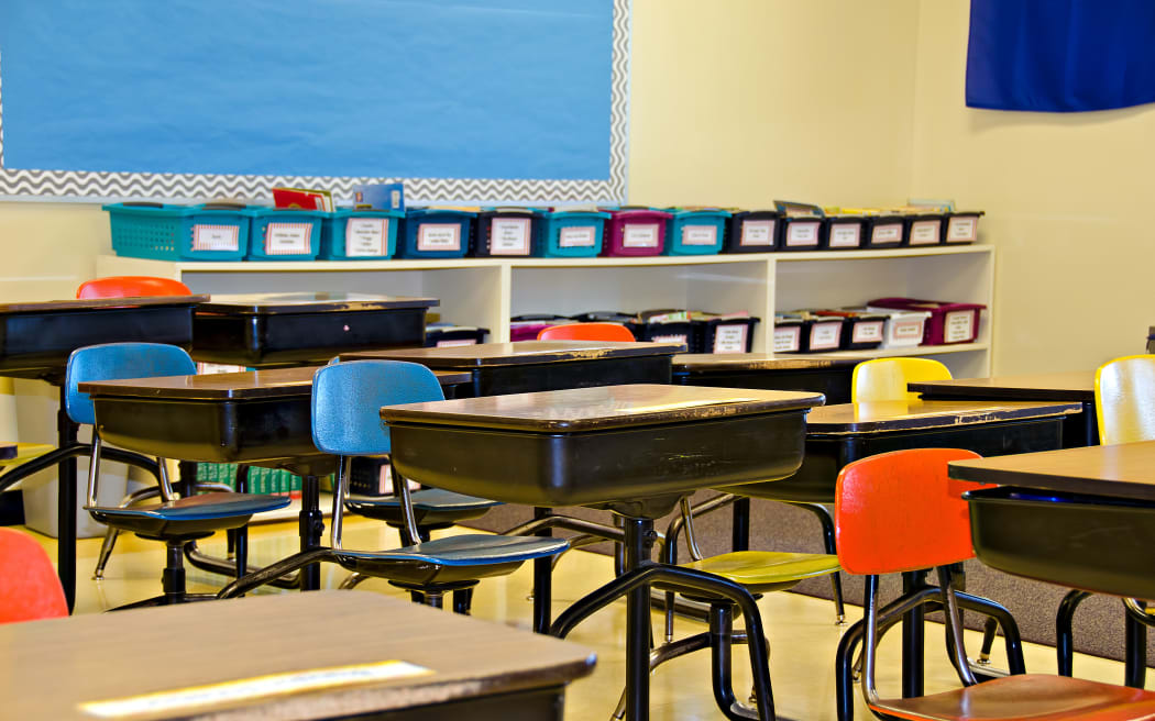 Students away from a classroom, which is filled with empty school desks.