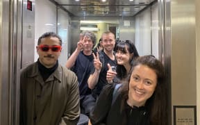 Five band members are squeezed into an elevator together with their gear, doing thumbs up and peace signs.