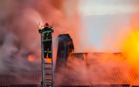 Firefighter or fireman on the ladder extinguishes burning fire flame with smoke on the apartment house roof.