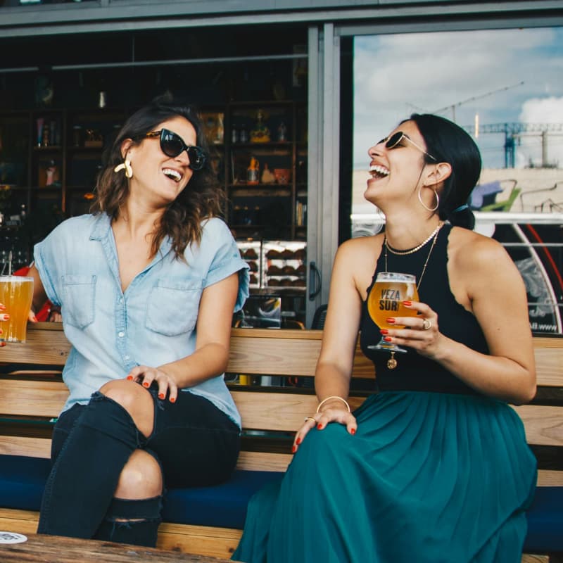 Two women in sunglasses sit outside at a bar laughing and drinking beer.