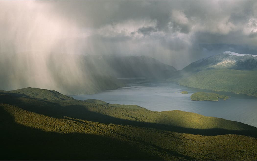 Snow showers blowing across Chalky Inlet in Fiordland National Park in the South Island of New Zealand. Forest covered hills in the foreground and snow capped mountains in the background.