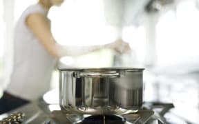Pot cooking on stove, woman in background (Photo by Laurence Mouton / AltoPress / PhotoAlto via AFP)
