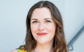 This is a headshot of Jessica Lockhart. She is looking towards the camera. Her brown hair frames her face. She has blue eyes and red lips drawn in a thin smile.
