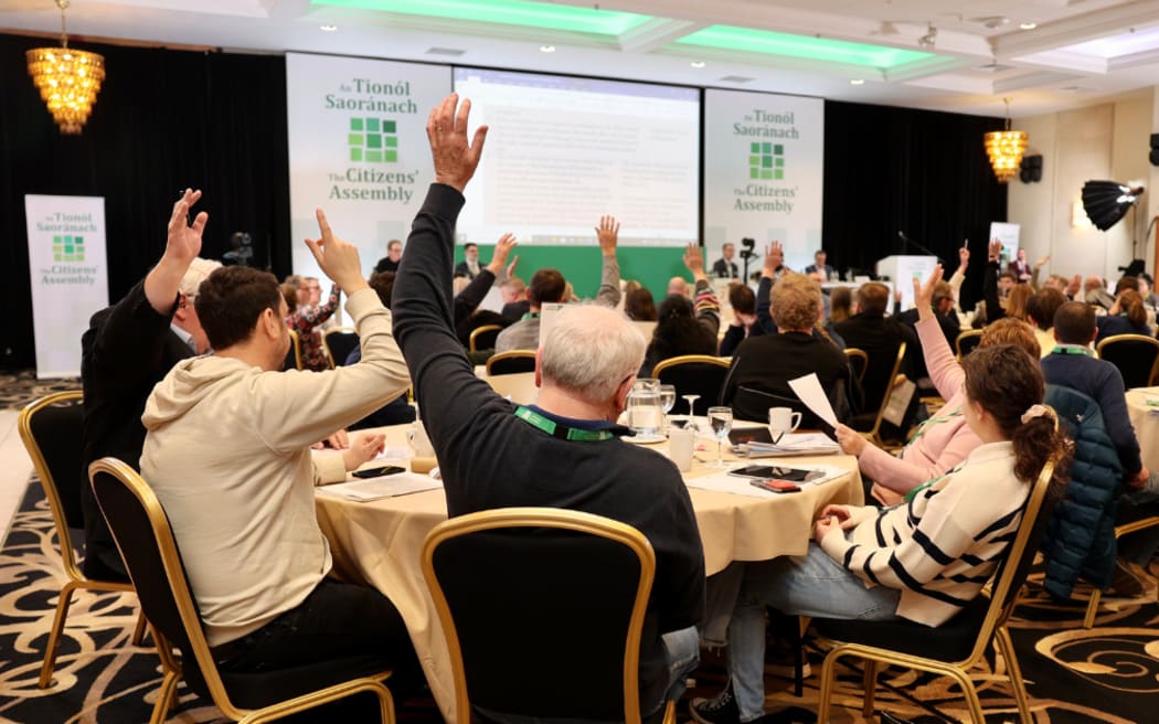 People in a conference room meeting as part of the Irish Citizens' Assembly. Seated in groups around tables, they appear to be voting on a resolution, with many people raising a hand.