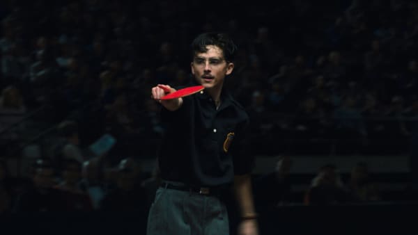 Timothee Chalamet, a young man with a moustache and glasses, holds a red ping-pong racquet as he points a finger.