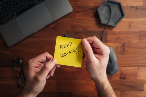 A pair of hands holding a yellow note with 'keep going!' written on it.