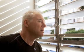 A man in his 60s looks out the window through open blinds. The man is wearing a black t-shirt. His hair gray. Head turned in profile. Glance tense. He looks at what's going on outside the window. Close-up shot.