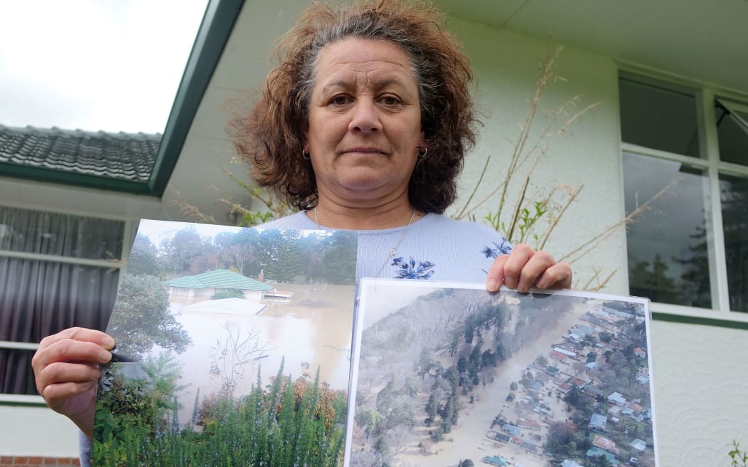 Woman stands in front of bungalow holding photos of flooded  home and surrounds in 2015