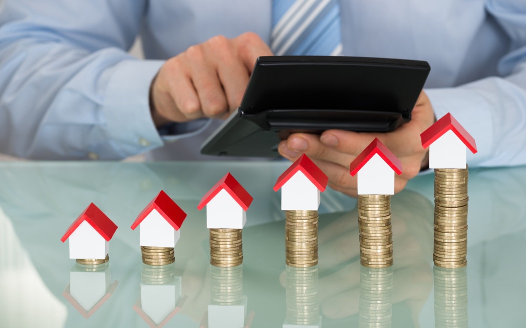 A man with a calculator inspects a line of houses stacked on top of gold coins.