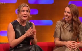 Kate Winslet speaks with clasped hands as Jacinda Ardern smiles and looks on.