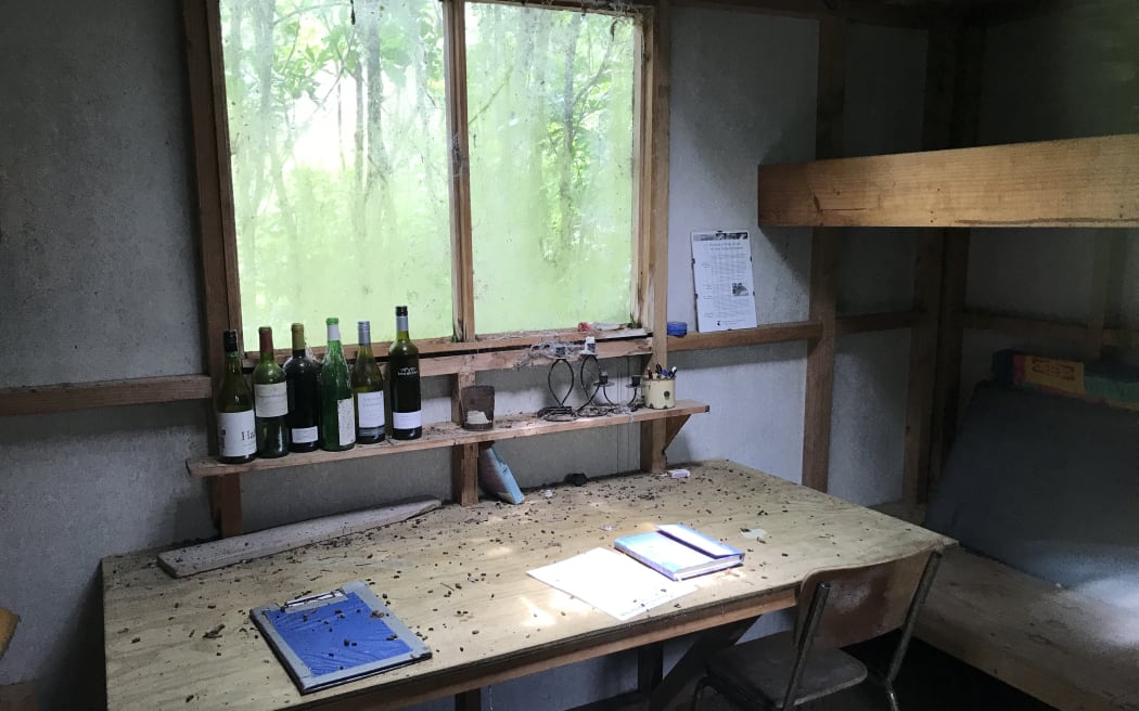 An old desk in a dimly lit room covered with dust. Greenery can be seen outside window, with a row of old wine bottles lining the windowsill.