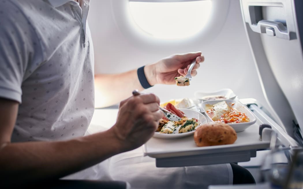Passenger on plane, holding a fork and eating from a meal in front of him on table, on a flight.