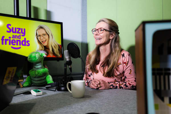 A woman sits at a desk with a microphone in front of her. Sitting on the table is a green dinosaur soft toy, with a TV screen behind it that says Suzy & Friends.