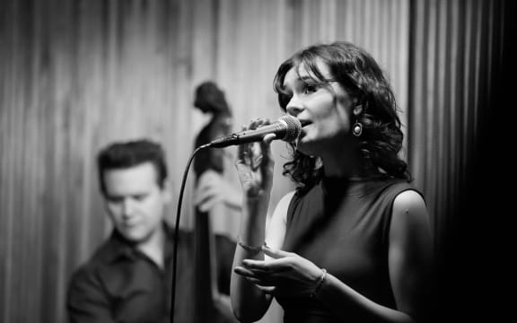 a black and white photo of ella singing and holding a microphone, with an out of focus double bass player in the background