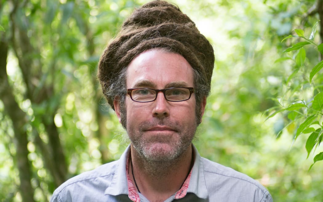 Duncan Kay is standing in front of some trees. it is a closeup head and shoulders shot.