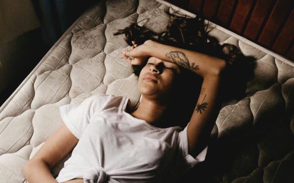 A woman reclines on an unmade bed, with one forearm across her forehead, the other on her stomach.