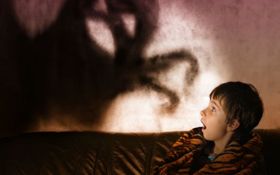 A photo of a boy in the dark with a shadow behind him in the shape of a monster with its arms extended.