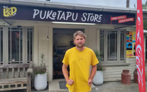 Puketapu Store owner Jaycen Maxwell stands outside the shop. He wears a yellow t-shirt.