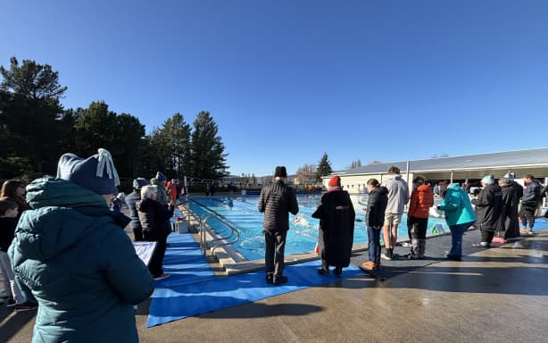 A sunny blue sky day at the outdoor pool in Alexandra, there are pine trees in the background. Around the pool are gathered a number of people dressed in warm jackets and hats. Some have clipboards and/or watches in hand.