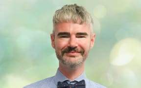 Scott stands in front of a soft green background. He wears a check shirt and a bowtie. He smiles.