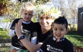 Amelia Kerr is wearing a Barnardos Aotearoa T-shirt and standing next to two smiling children.