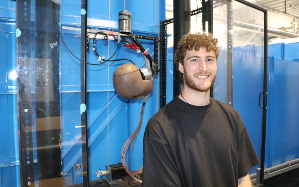 A midshot of a man in a black t-shirt. He has short curly brown hair and facial hair. He is standing in front of a head-shaped contraption.