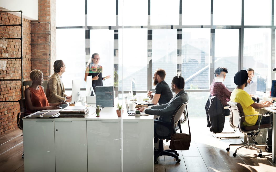 Office workers sitting at their desks in a brightly lit office with floor to ceiling windows letting in the sun.