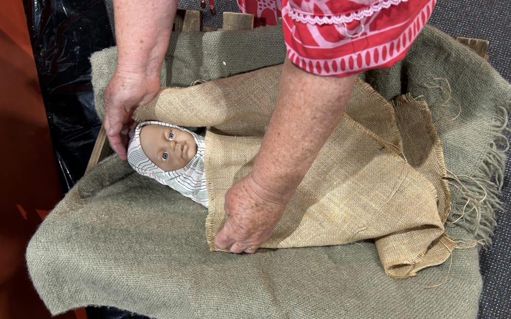 Photo of a doll being wrapped in baby cloth, with a pair of hands from a volunteer tucking the baby underneath a linen blanket.