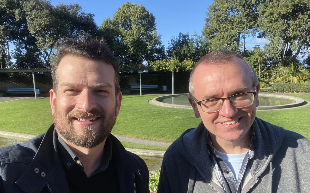 Rob and Chris smile at the camera. They're standing in a park.