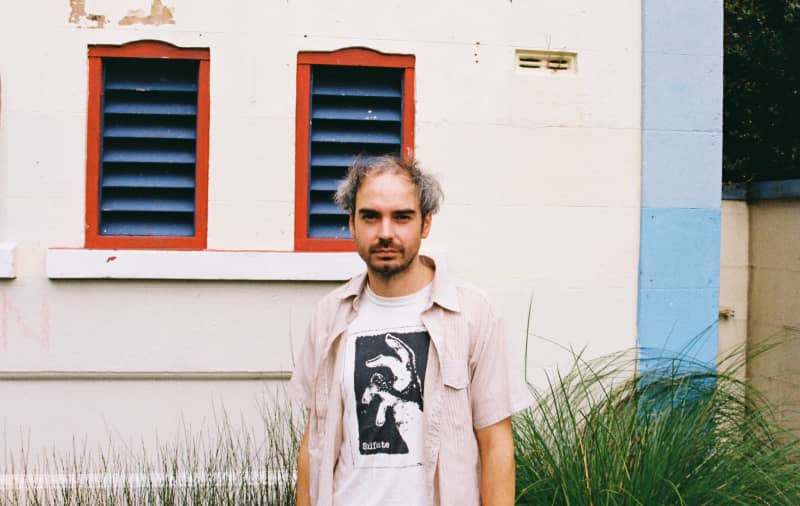 Peter McCall stands in front of a building wearing a beige short sleeved shirt over a white t=shirt with a black graphic print.